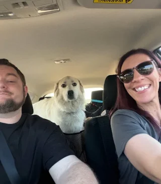 A woman and man in the front seat of a car with a white dog looking at the camera.